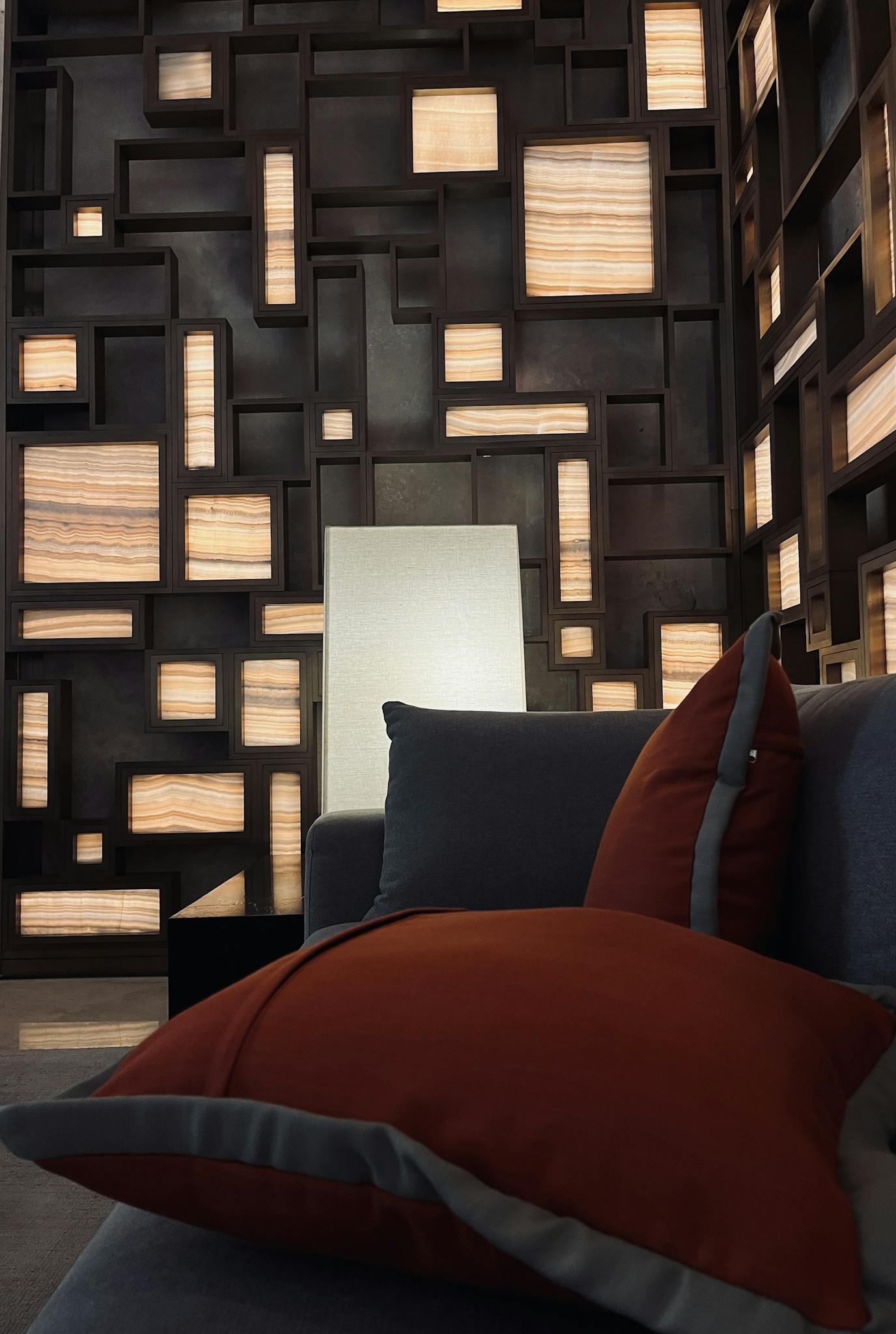 Contemporary living room with a geometric backlit wall, orange pillows, and grey couch.